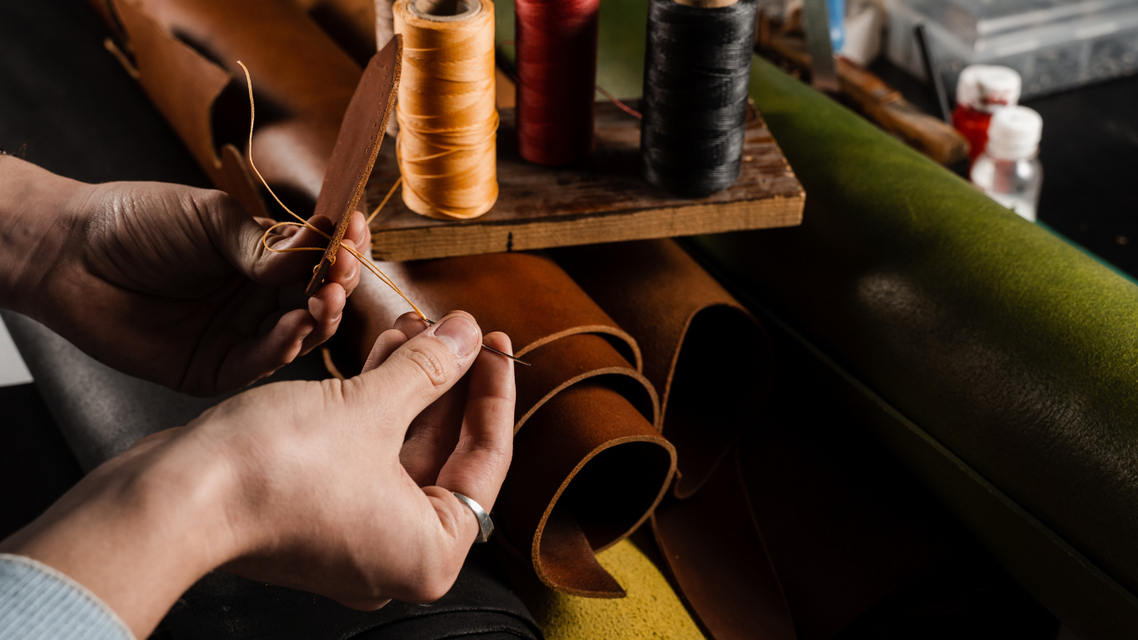 Hands sewing leather straps with colored thread spools on a workbench