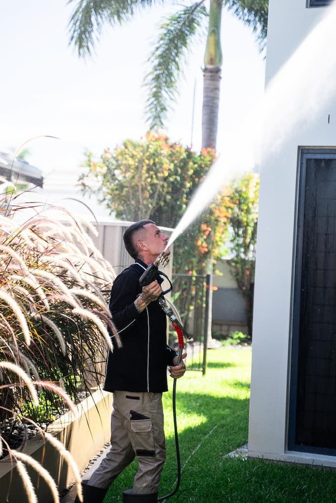 Man spraying water on a building. He is outside, holding a hose, aiming upward with a focused expression.
