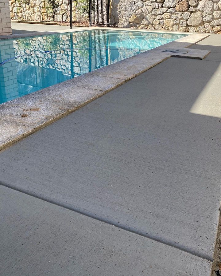 A pool with a concrete surround. Blue water reflects sky. A stone wall is in the background.