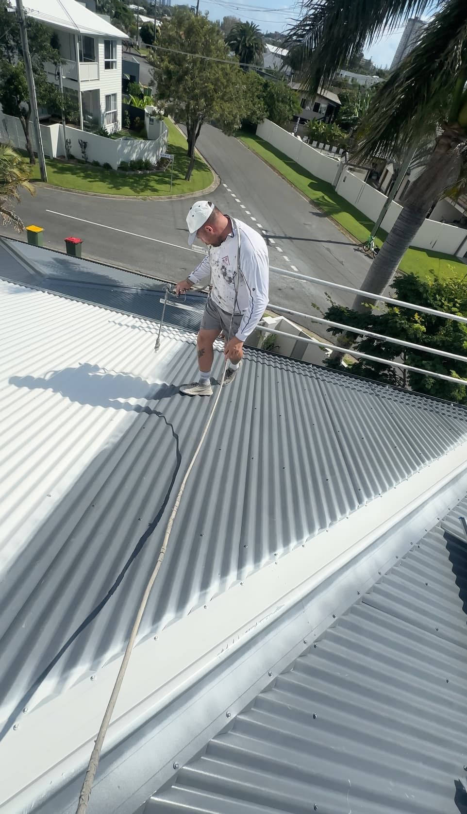 Man painting a corrugated metal roof white, using a roller. Safety rope attached.