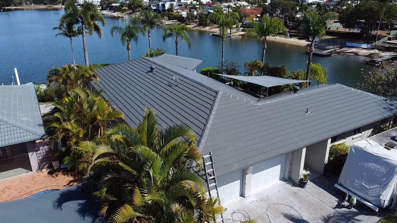 A house with a gray roof sits near a body of water with palm trees in the background.