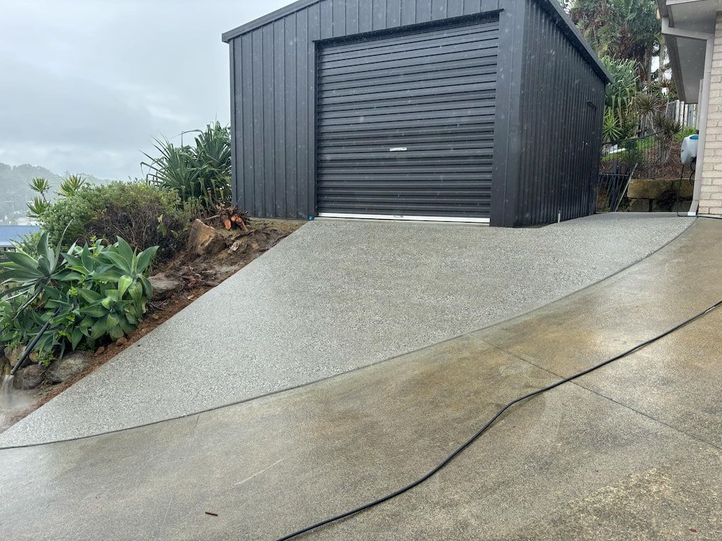 Black shed with a roller door at the end of a concrete driveway; overcast day.