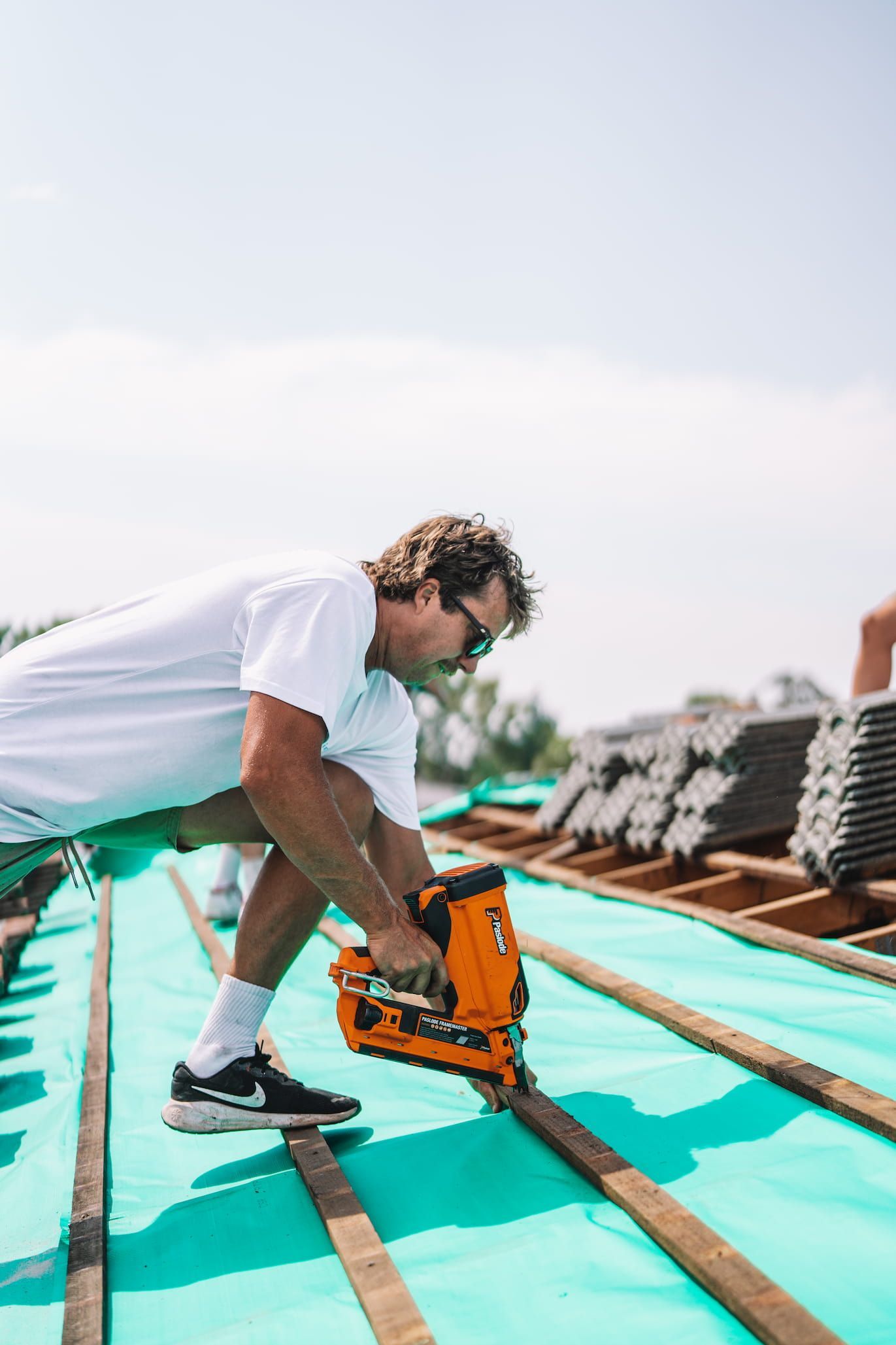 Man on a roof using a nail gun; installing roofing material on a sunny day.