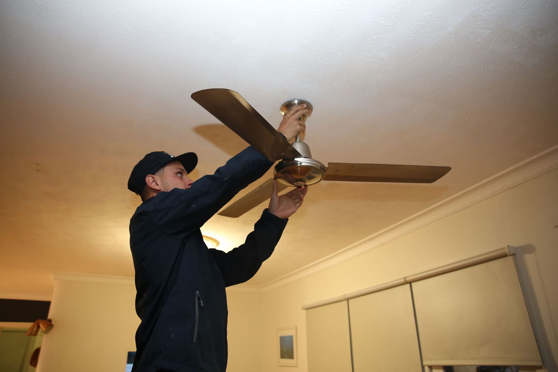 Man in dark clothes installing a ceiling fan in a room with white walls.