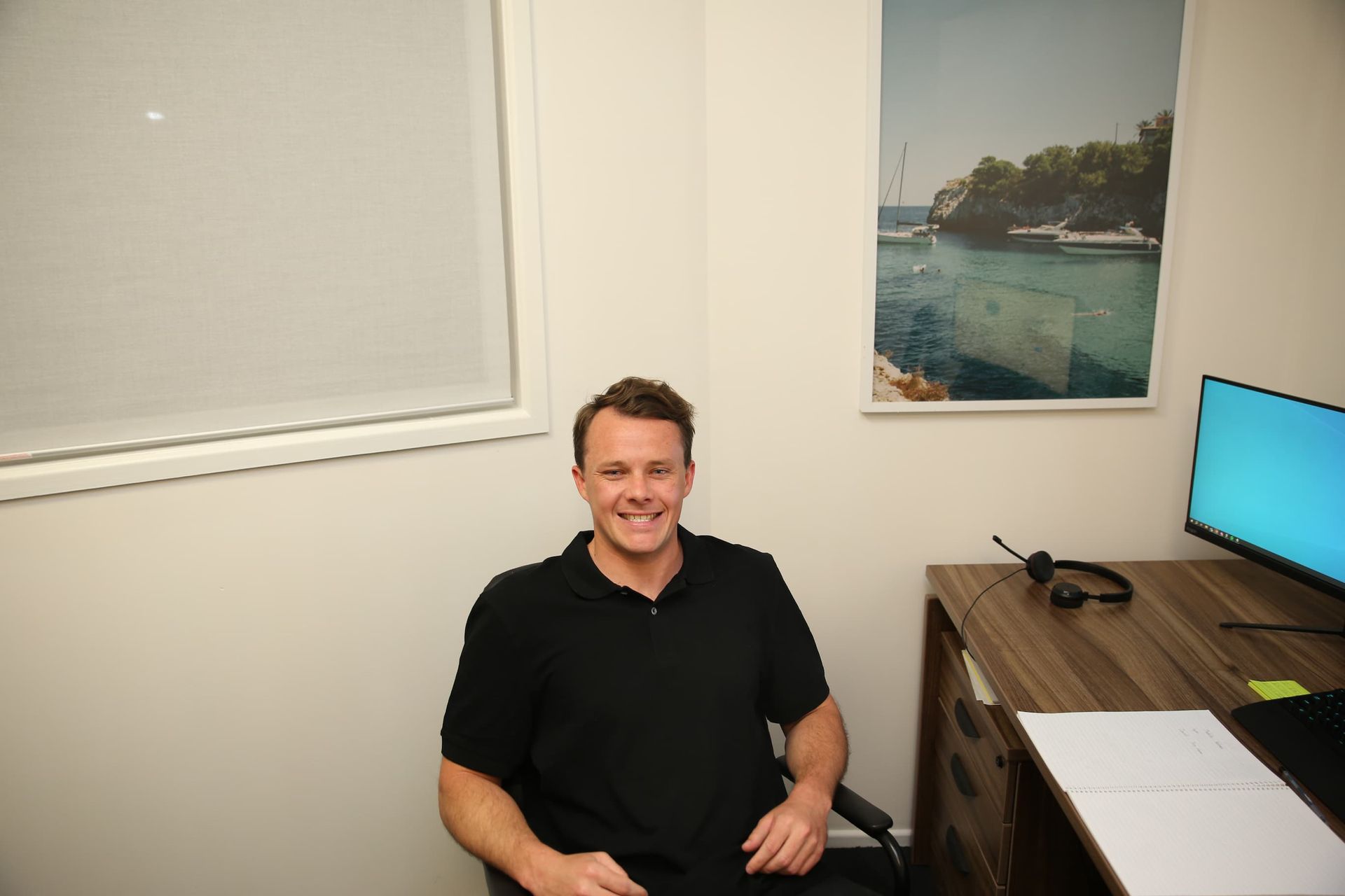 Man smiling in a small office with a window, desk, and seascape artwork.