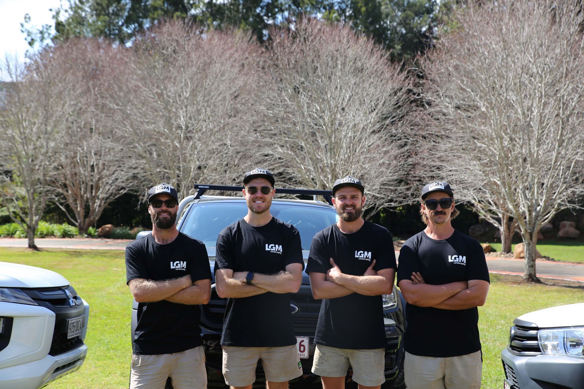 Four men in black shirts and hats stand in front of a black SUV and two white trucks in a park.