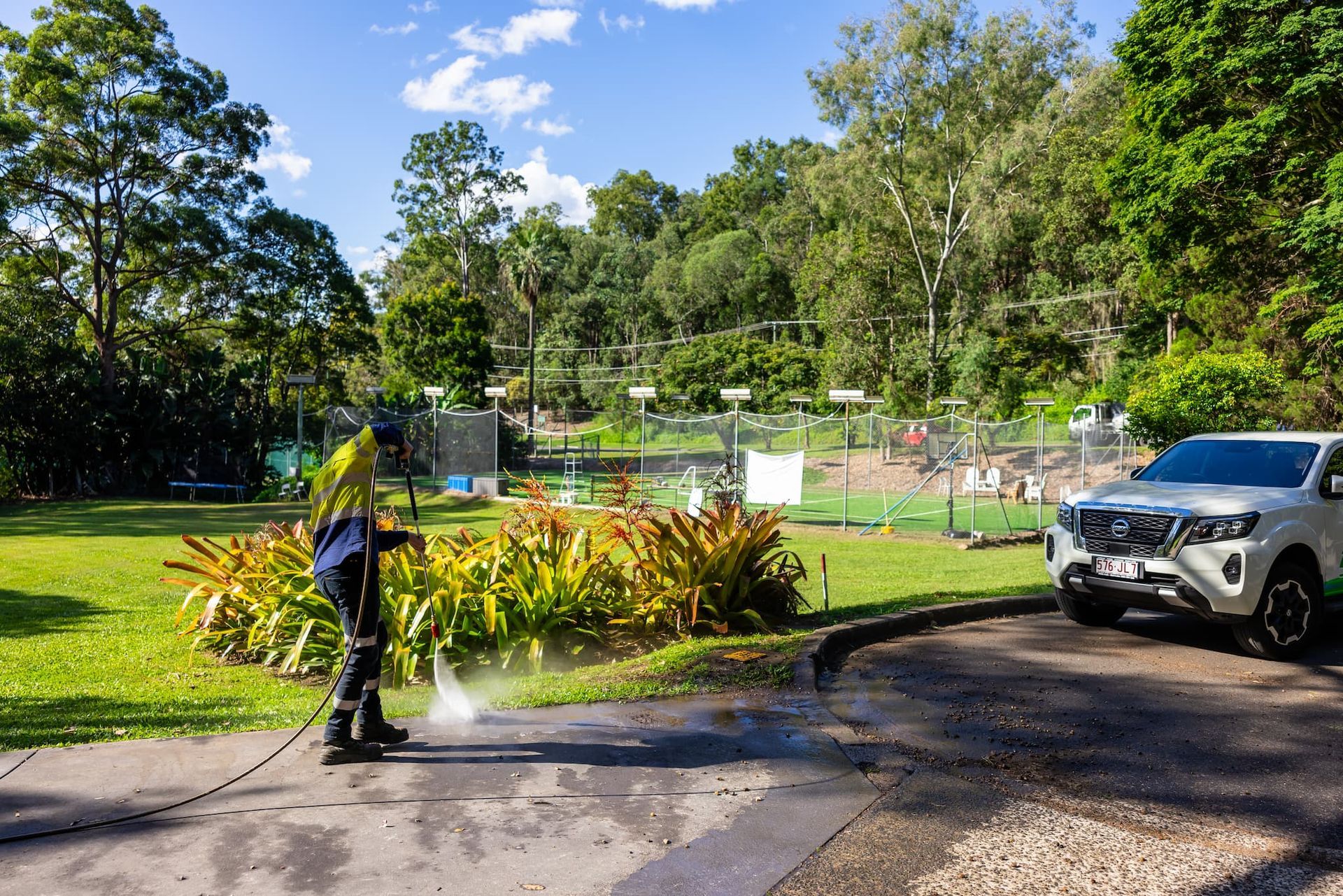 Person pressure washing a driveway near a green space and white car.