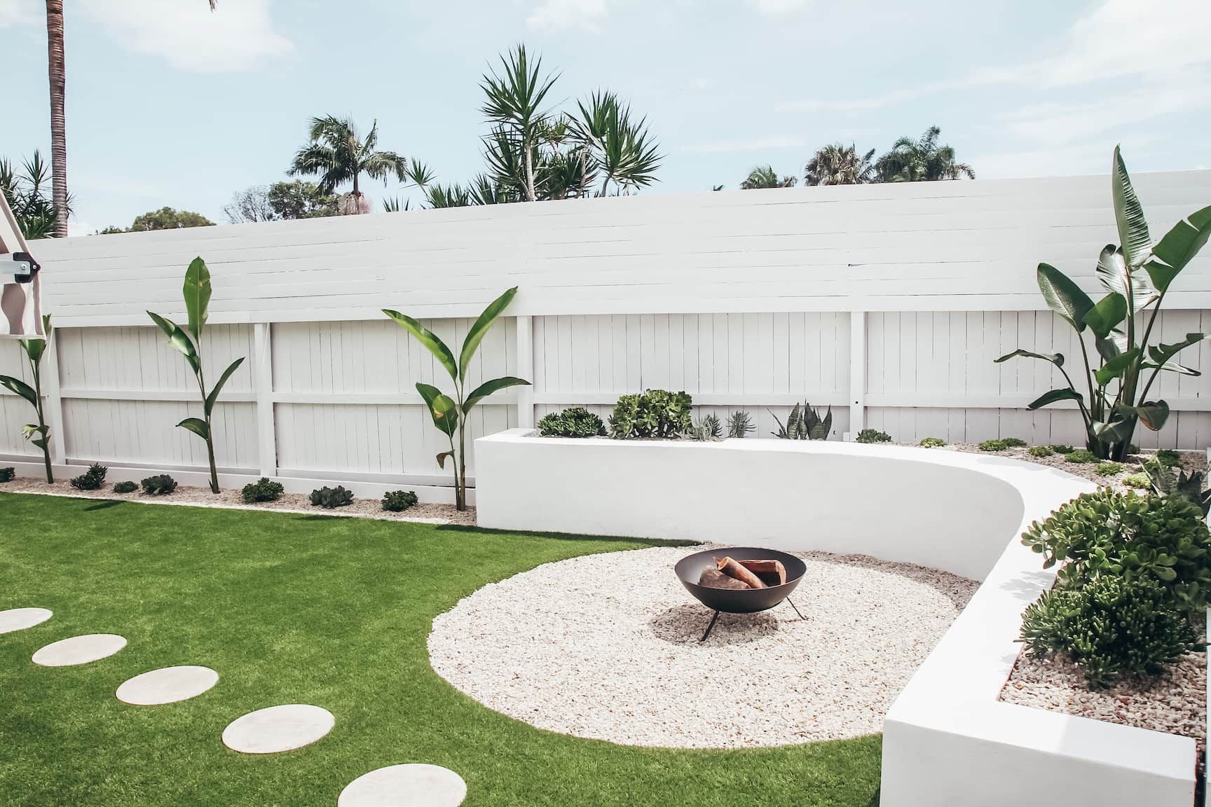 A backyard with a white fence, green grass, a fire pit area with white stones, and plants.