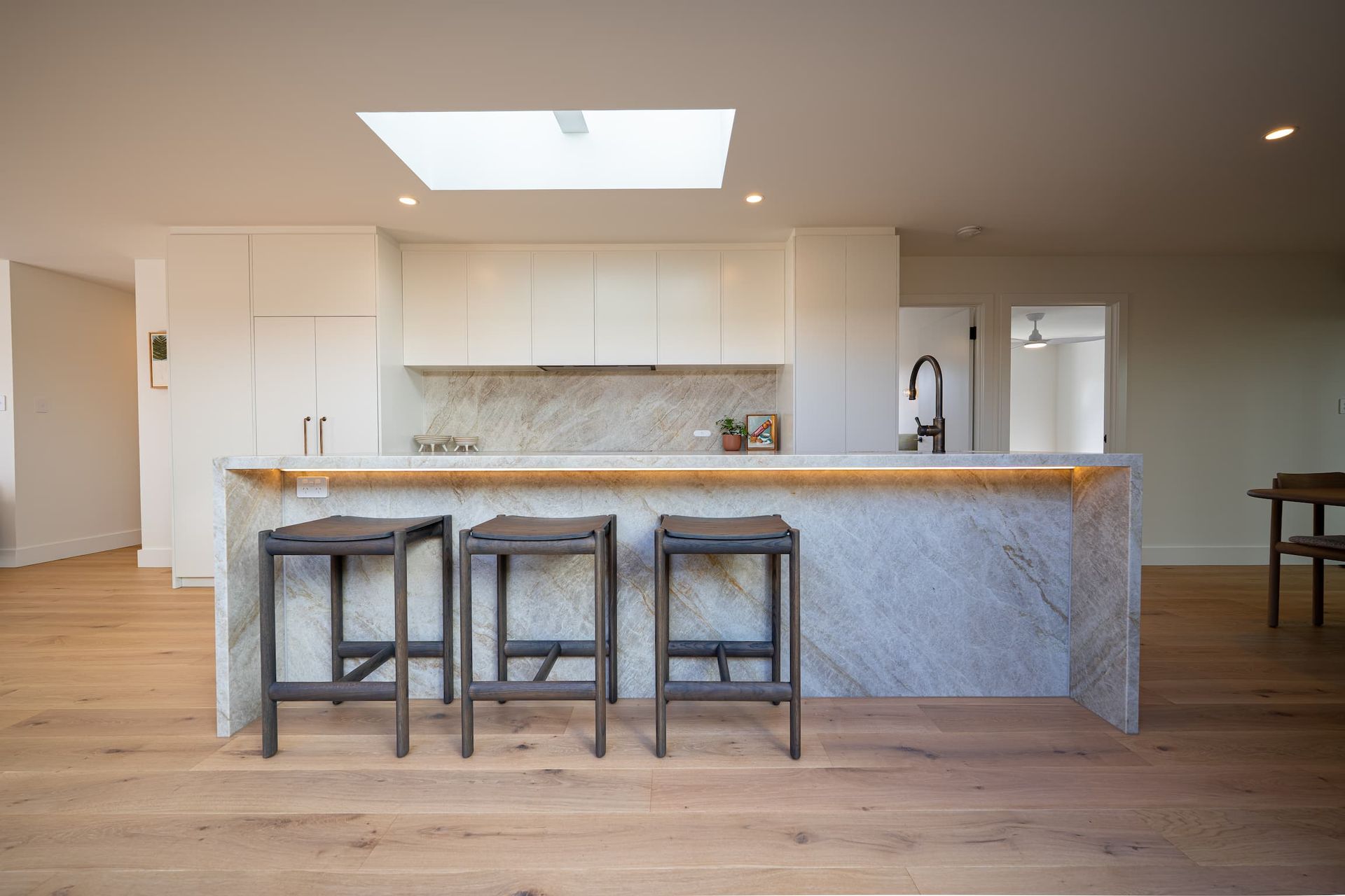 Kitchen with marble island, wood flooring, three stools, and white cabinetry. Skylight overhead.