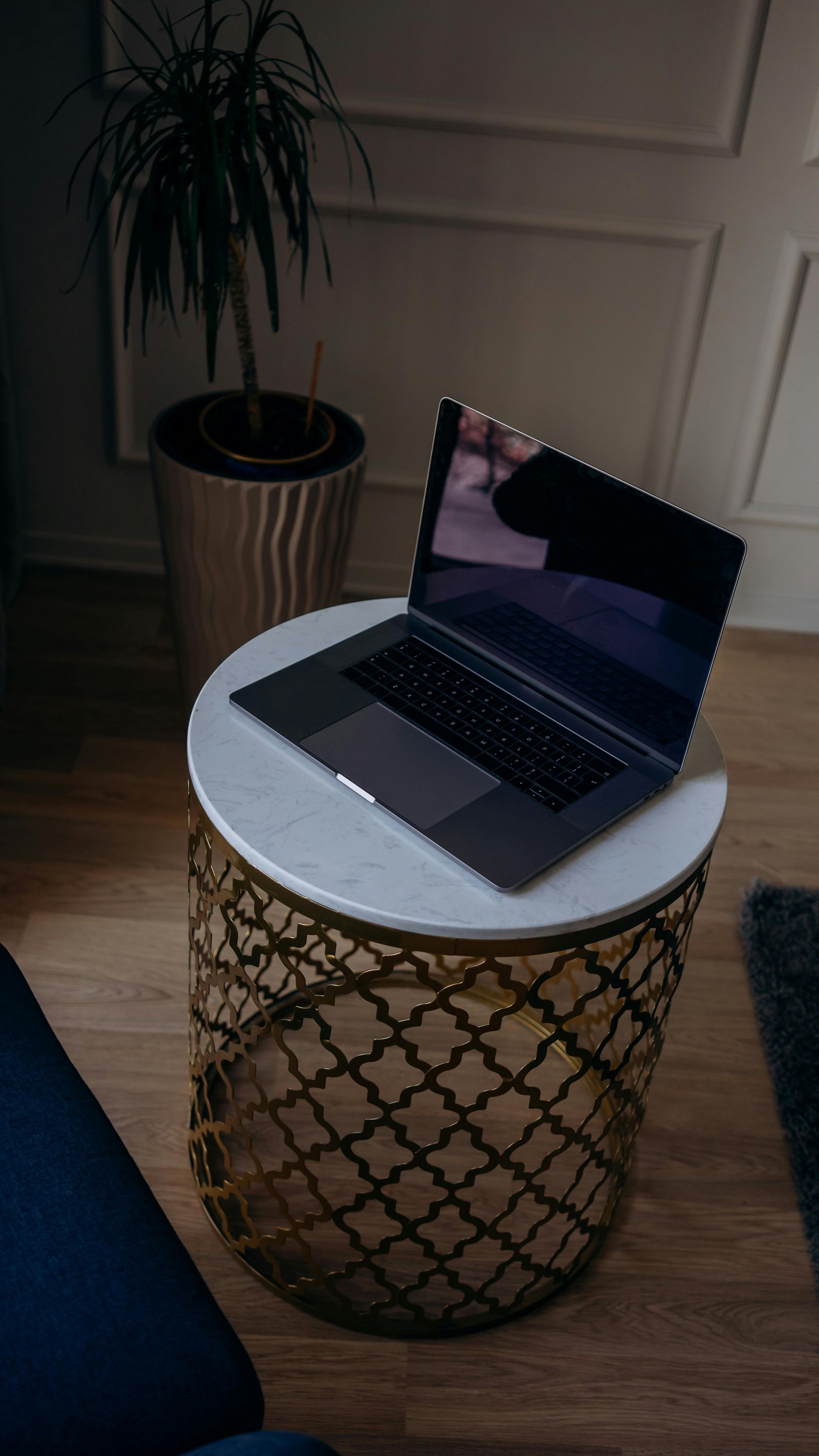 Open laptop on a small round table in a dim room, with a potted plant nearby.