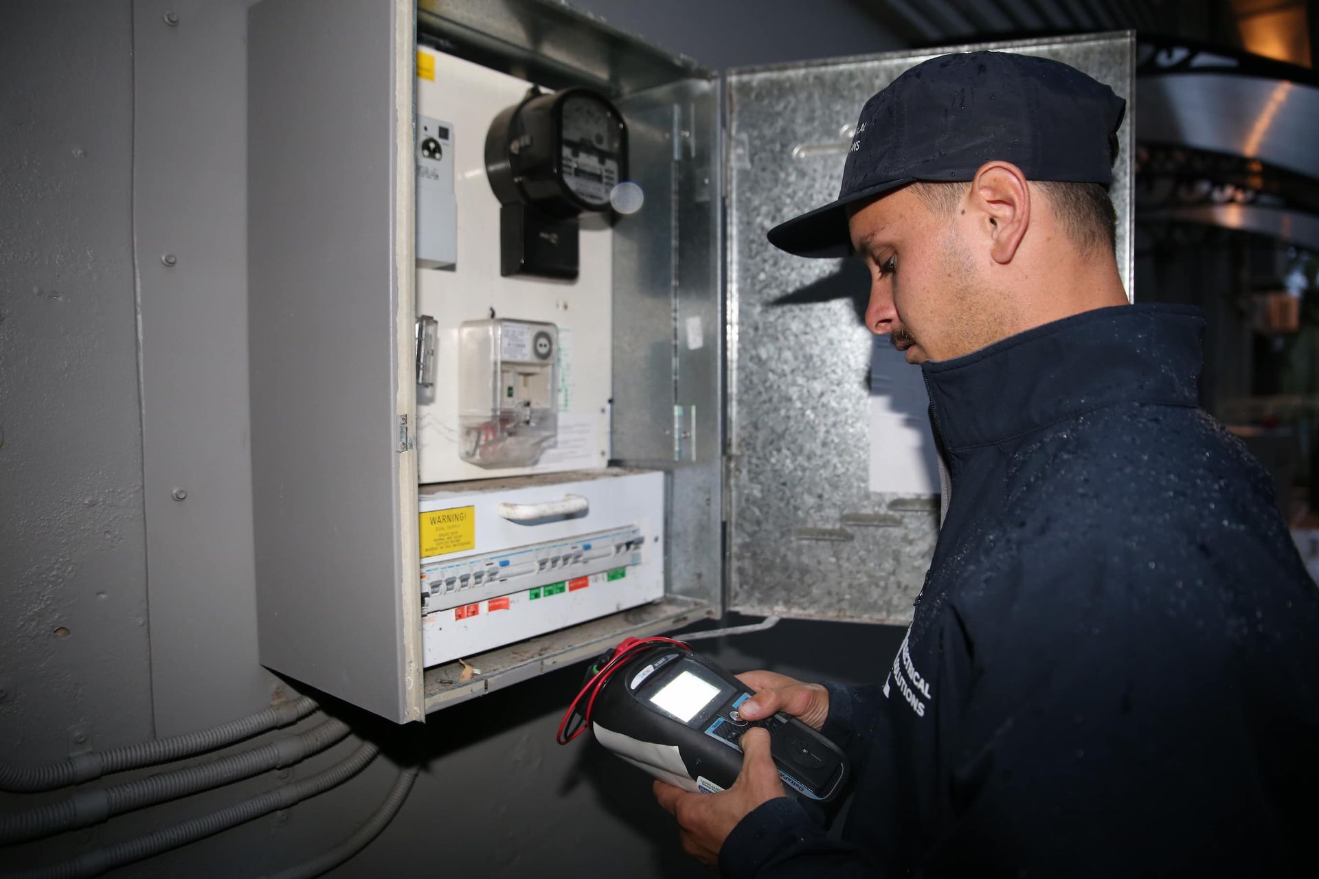 Electrician in a dark room checking electrical panel with a handheld device.