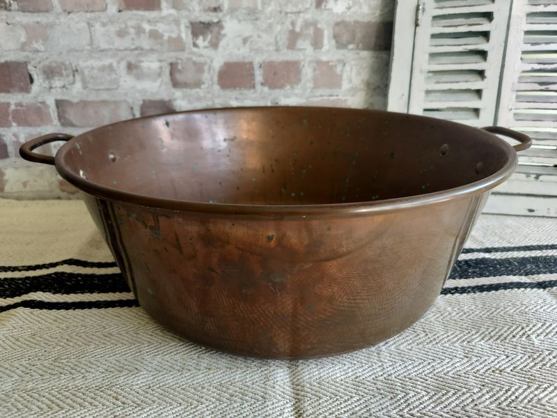 A large copper bowl with handles is sitting on a rug in front of a brick wall.