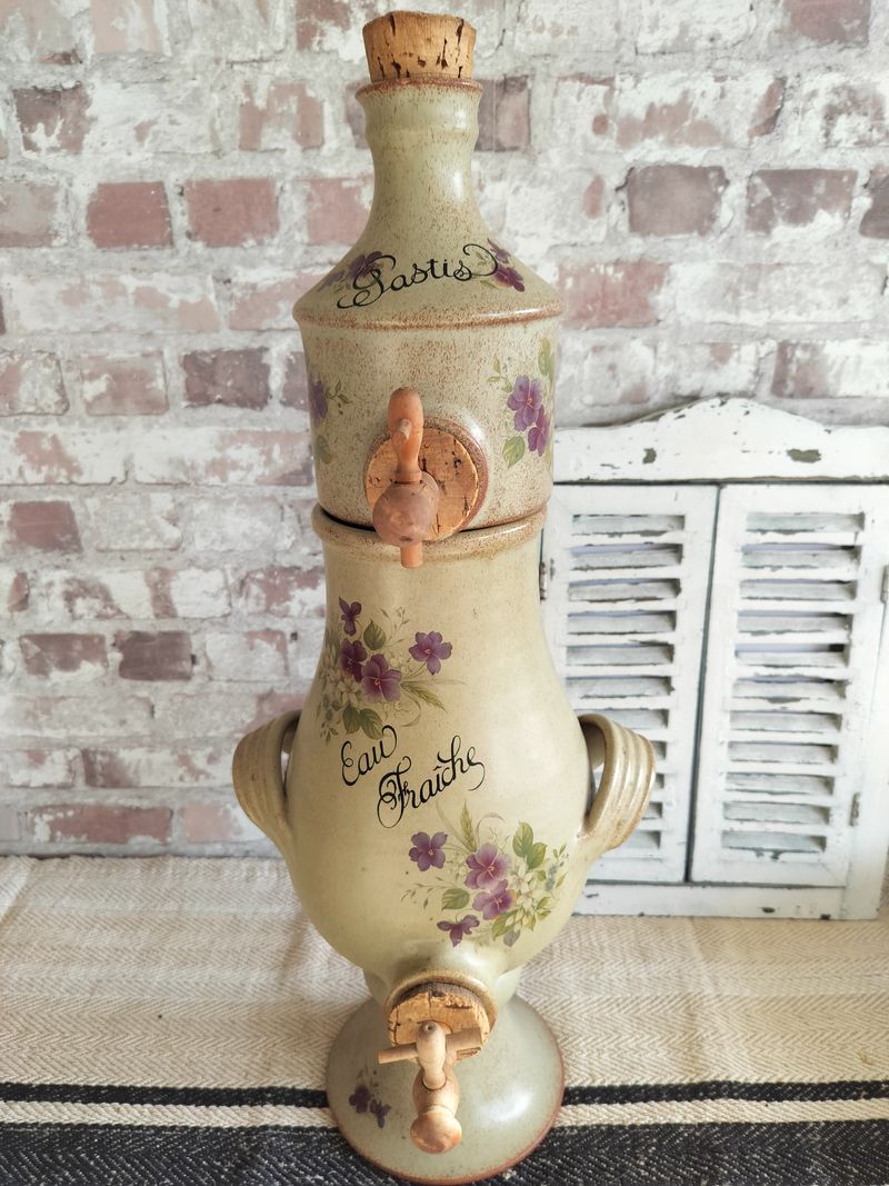 A ceramic bottle with purple flowers on it is sitting on a rug in front of a brick wall.