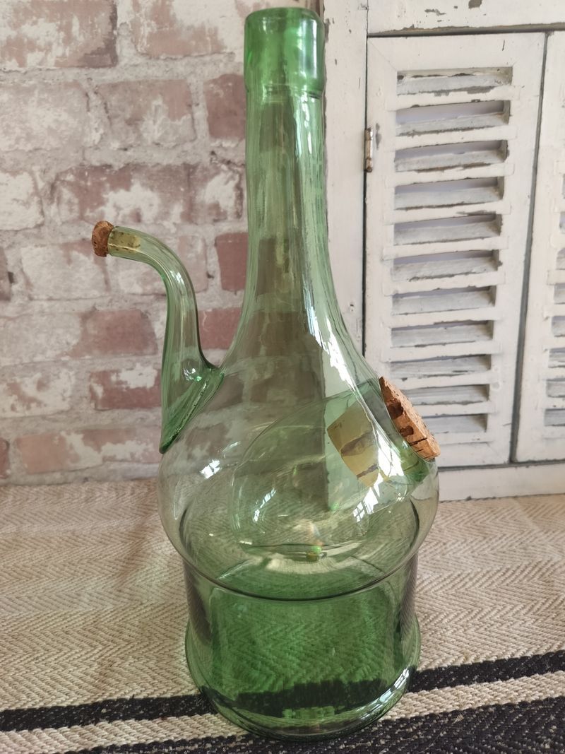 A green glass bottle with a handle is sitting on a rug in front of a brick wall.