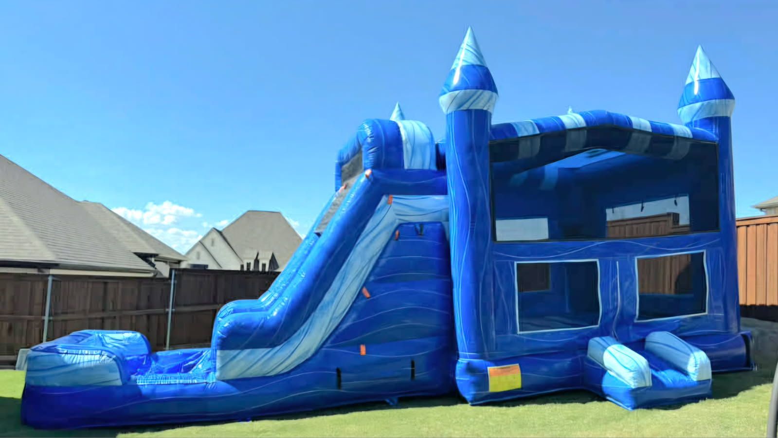 A blue and white inflatable bounce house with an attached slide, situated on a grassy lawn in a suburban backyard.