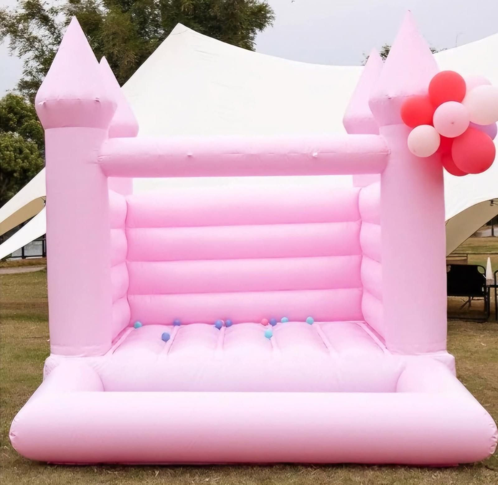 A light pink inflatable bounce house with a castle design, accented by a small cluster of balloons, set on grass.