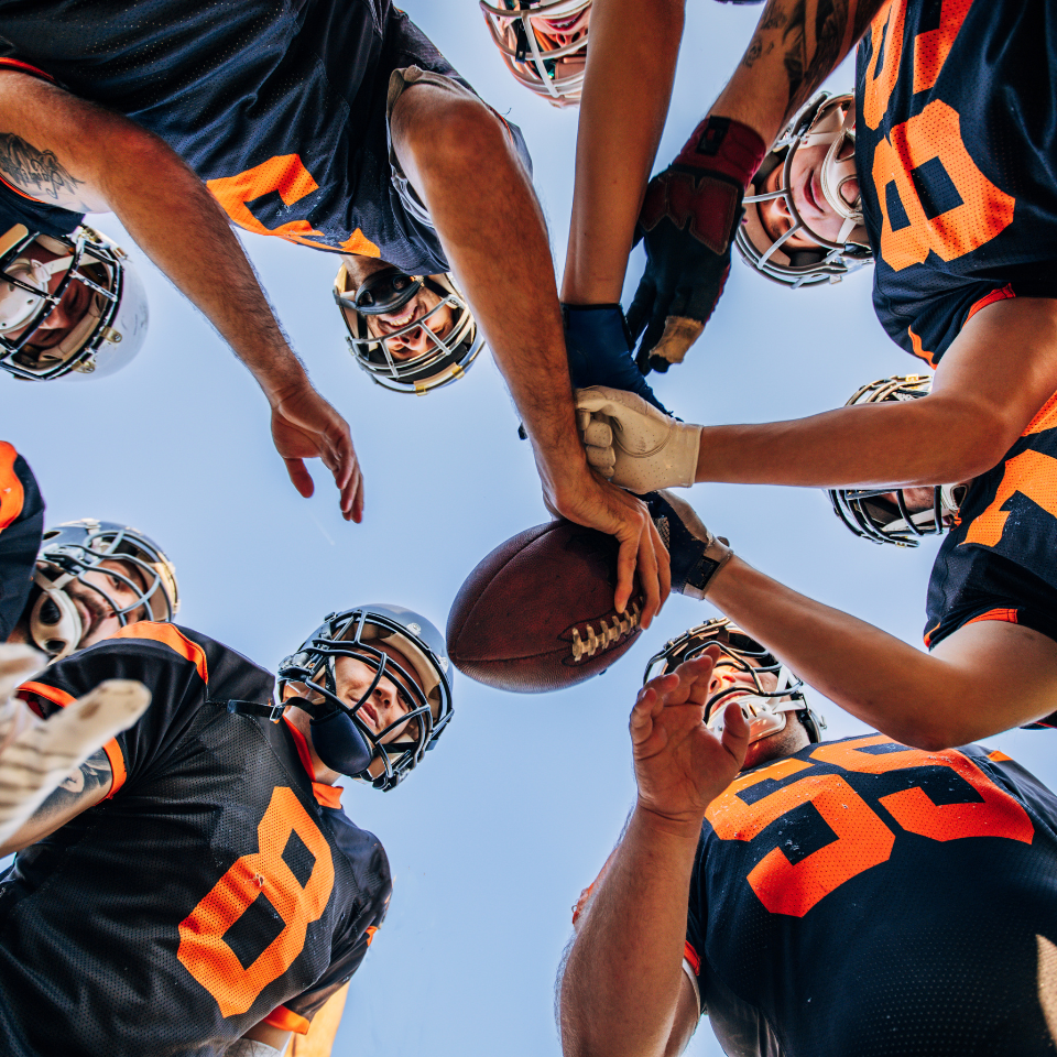 Football players huddle around a football, reaching hands together, blue sky backdrop.