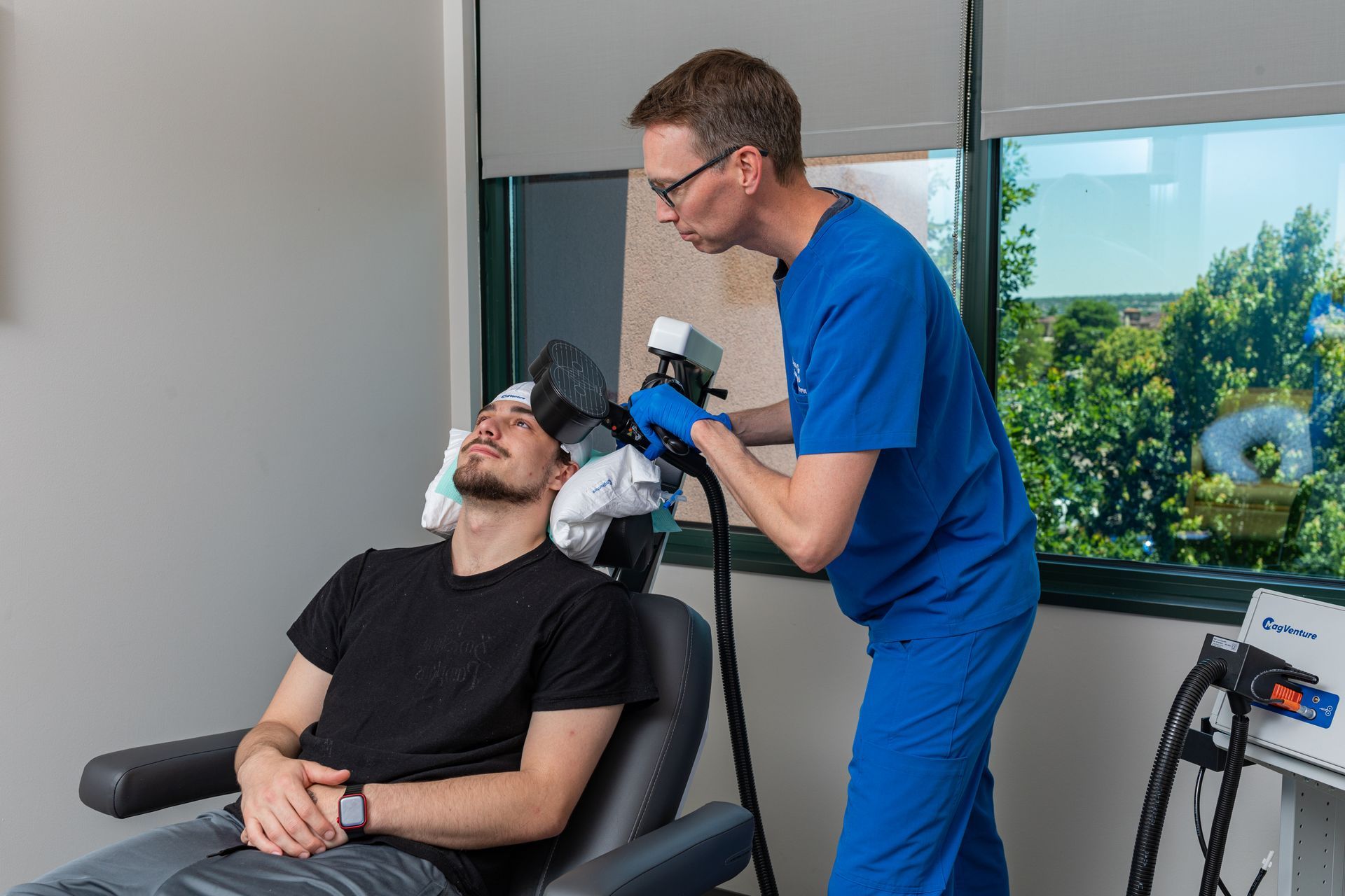 Woman in blue scrubs using a device on a patient's head. White towel, window with green trees.