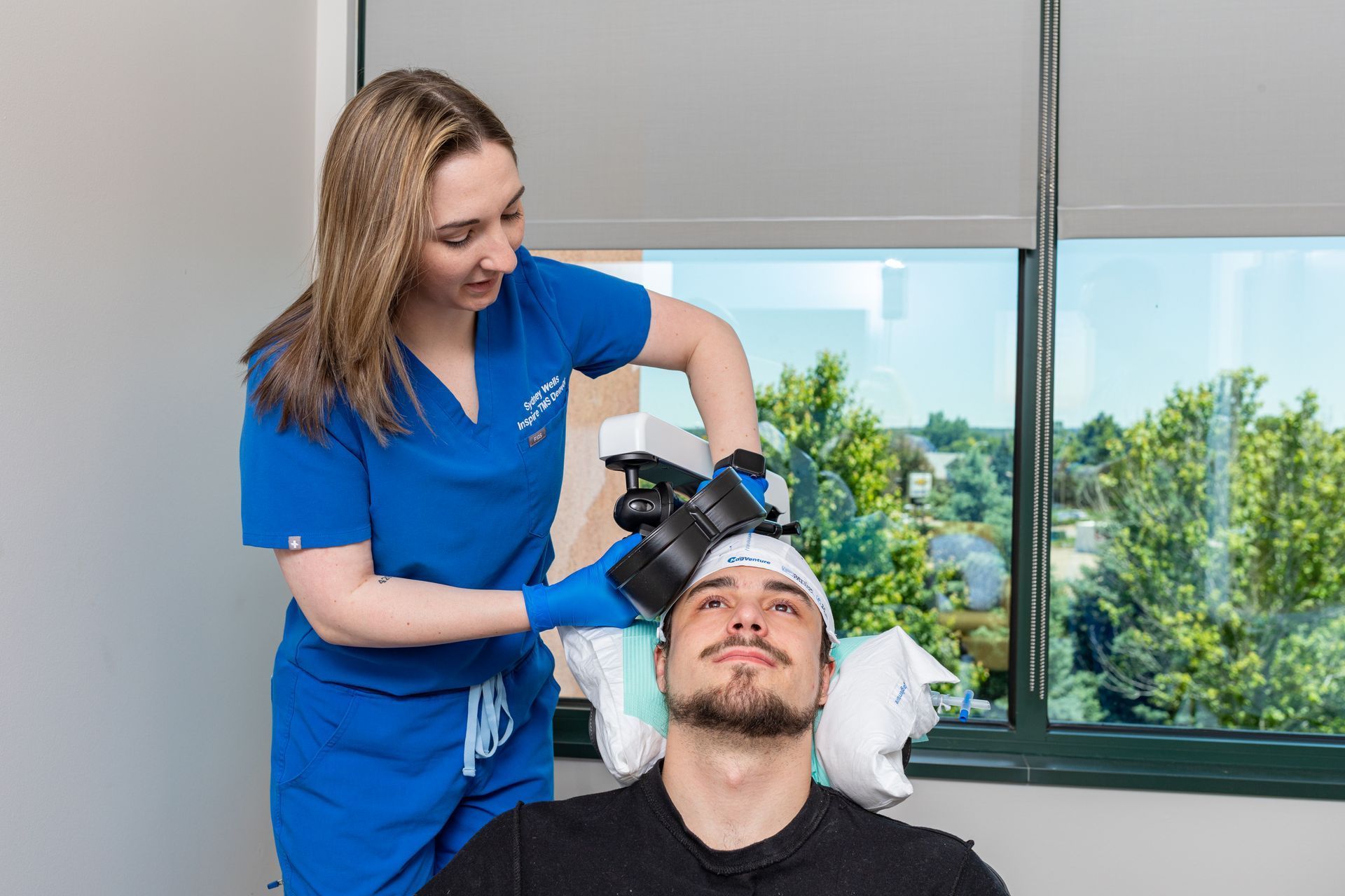 A medical professional in blue scrubs places a device on a patient's head. Inside a medical office by a window.