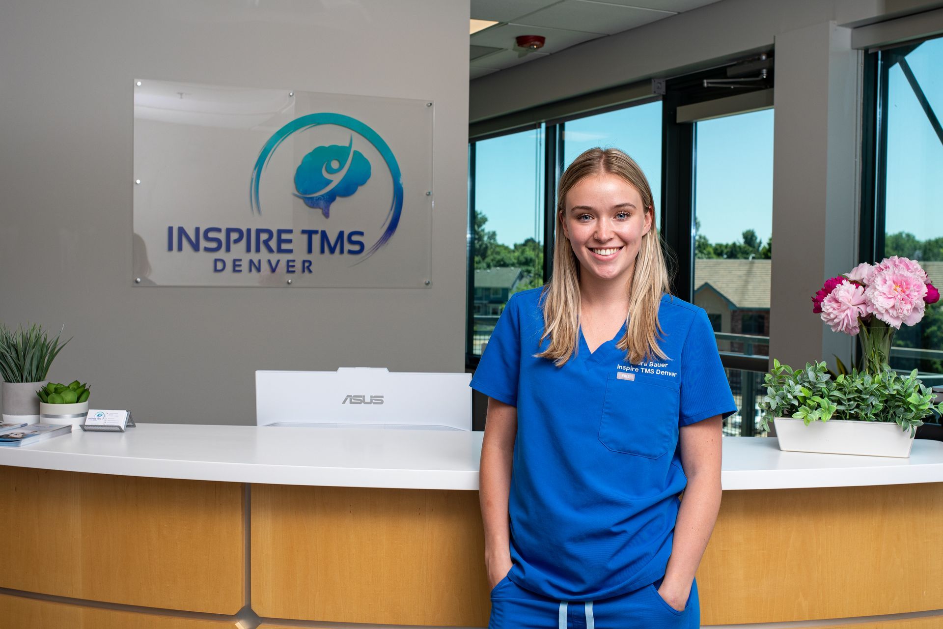 Man at desk smiles while talking to a person seated in front of him. A window with trees in the background.