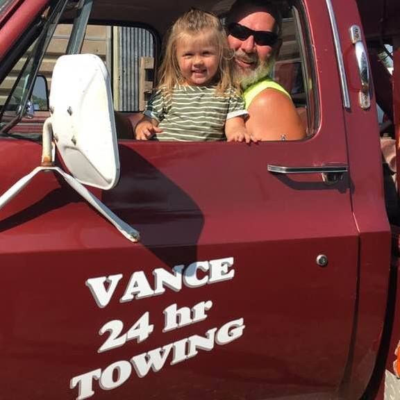 Man and young child in a red tow truck, smiling. Text on door: Vance 24hr Towing.