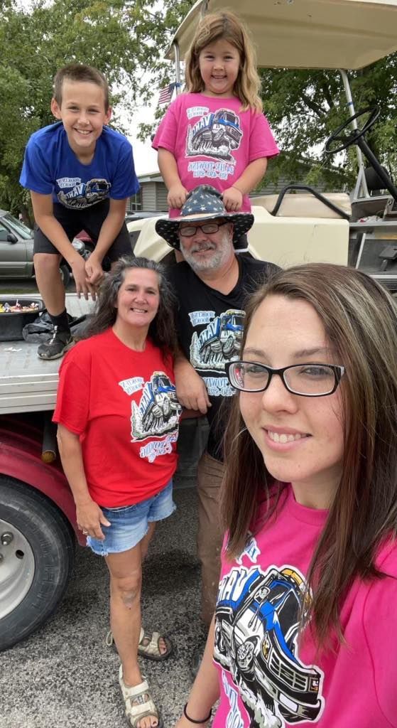 Family posing in front of golf cart. Wearing matching shirts, boy on top, girl behind, adults in front, outdoor setting.