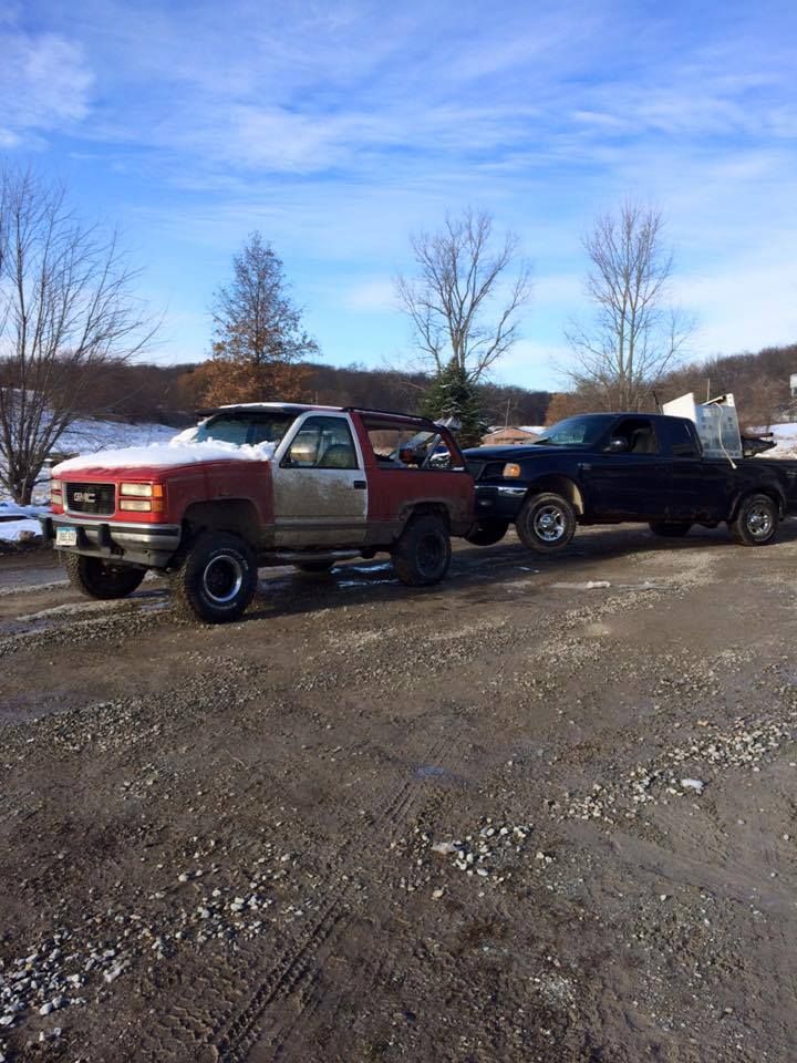 Two pickup trucks; a red and silver GMC pulling a blue Ford on a dirt road, under a blue sky.