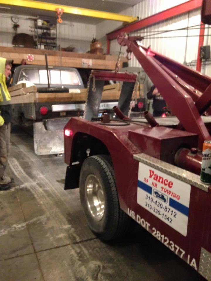 Tow truck in a garage, lifting a truck bed. Red truck, wooden beams, overhead crane, and a worker visible.