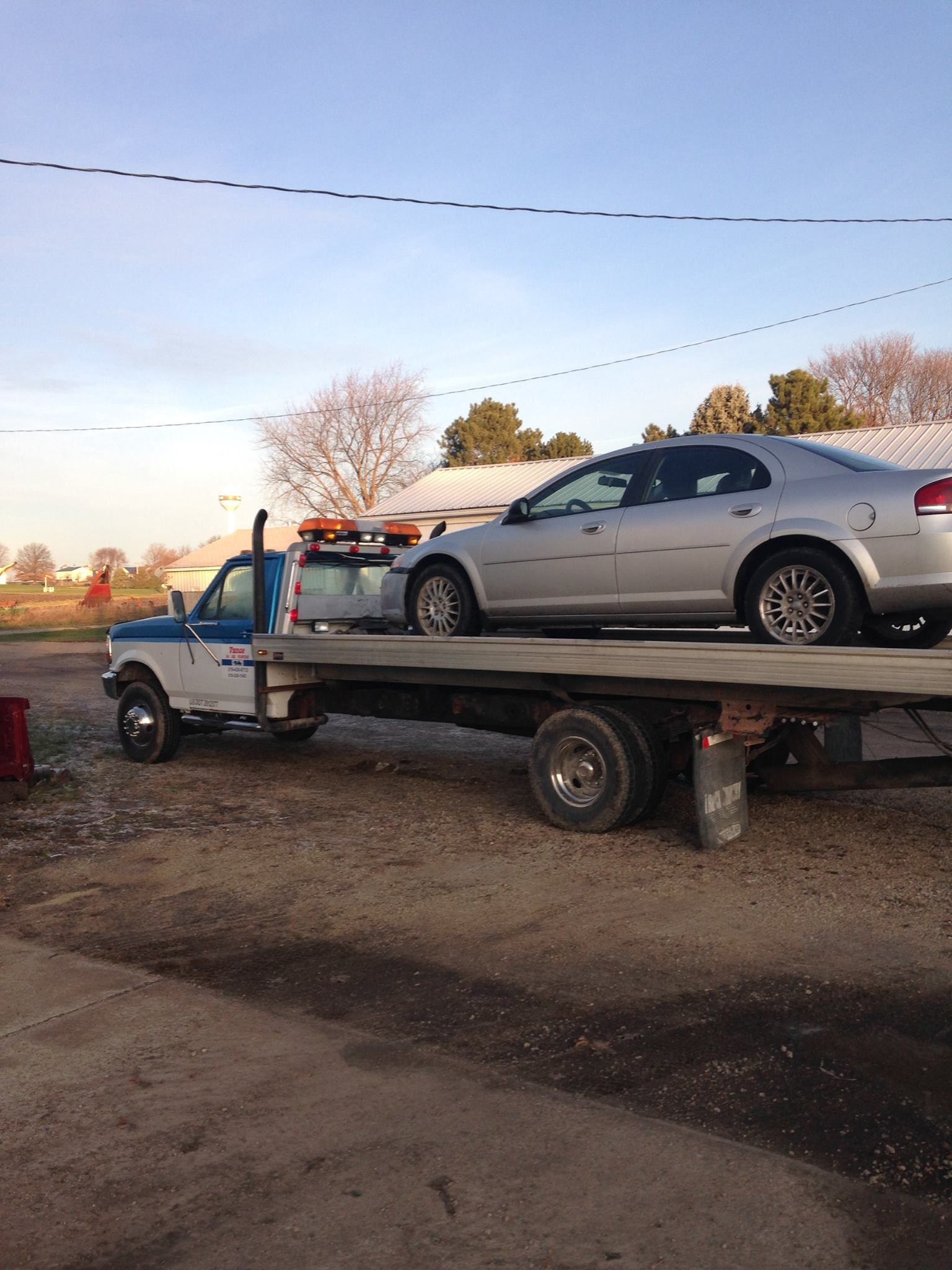 A silver sedan being towed on a flatbed tow truck on a gravel lot under a blue sky.