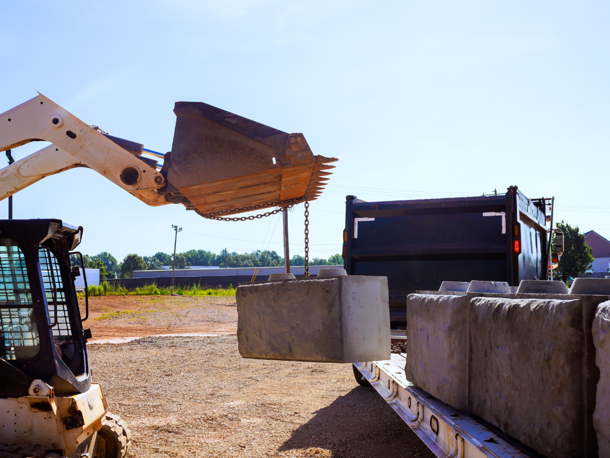 Skid steer loading a large concrete block onto a trailer with a dump truck in the background on a sunny day.