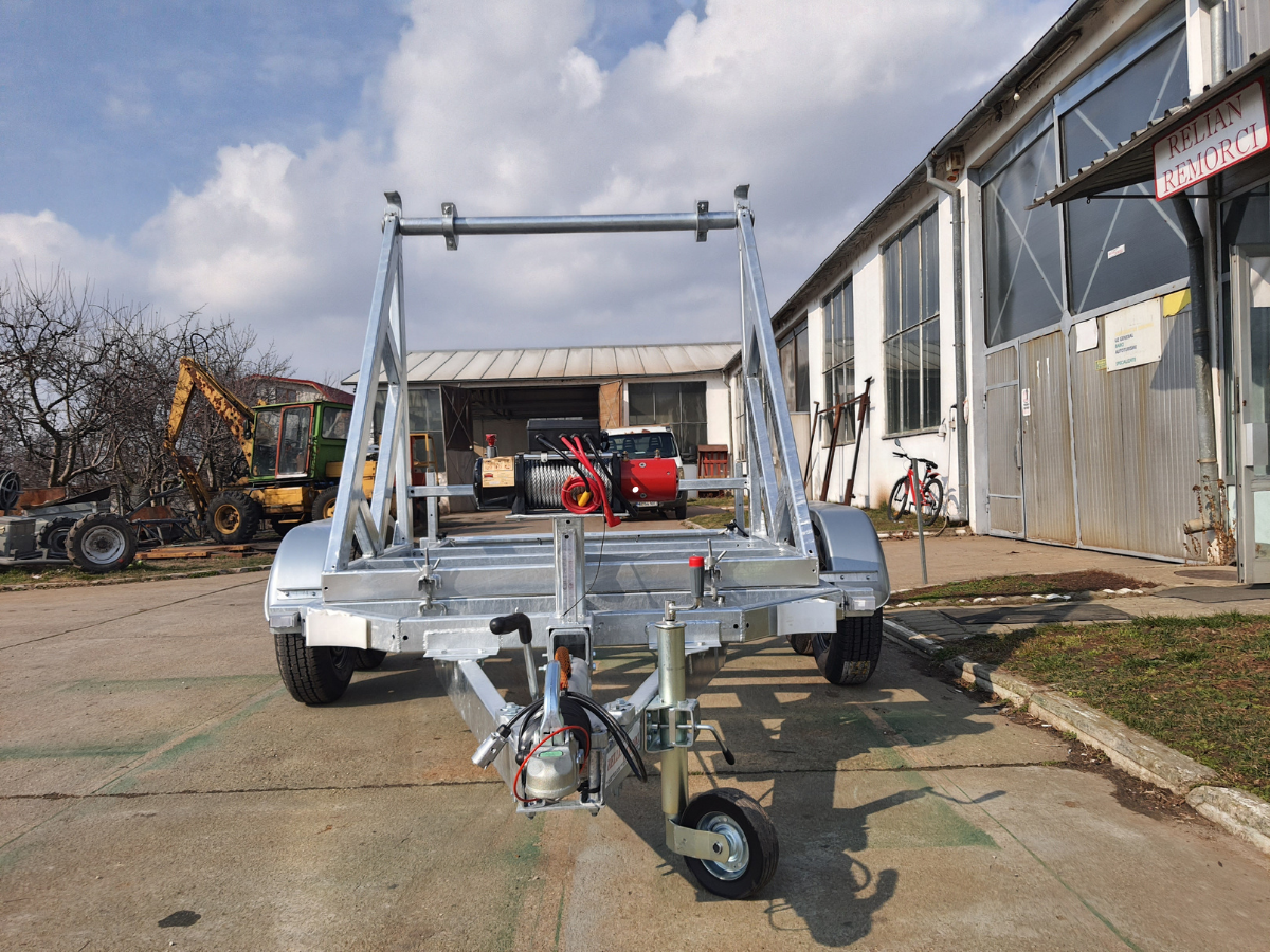 Galvanized steel cable reel trailer on a paved surface, parked near a building with other vehicles in the background.