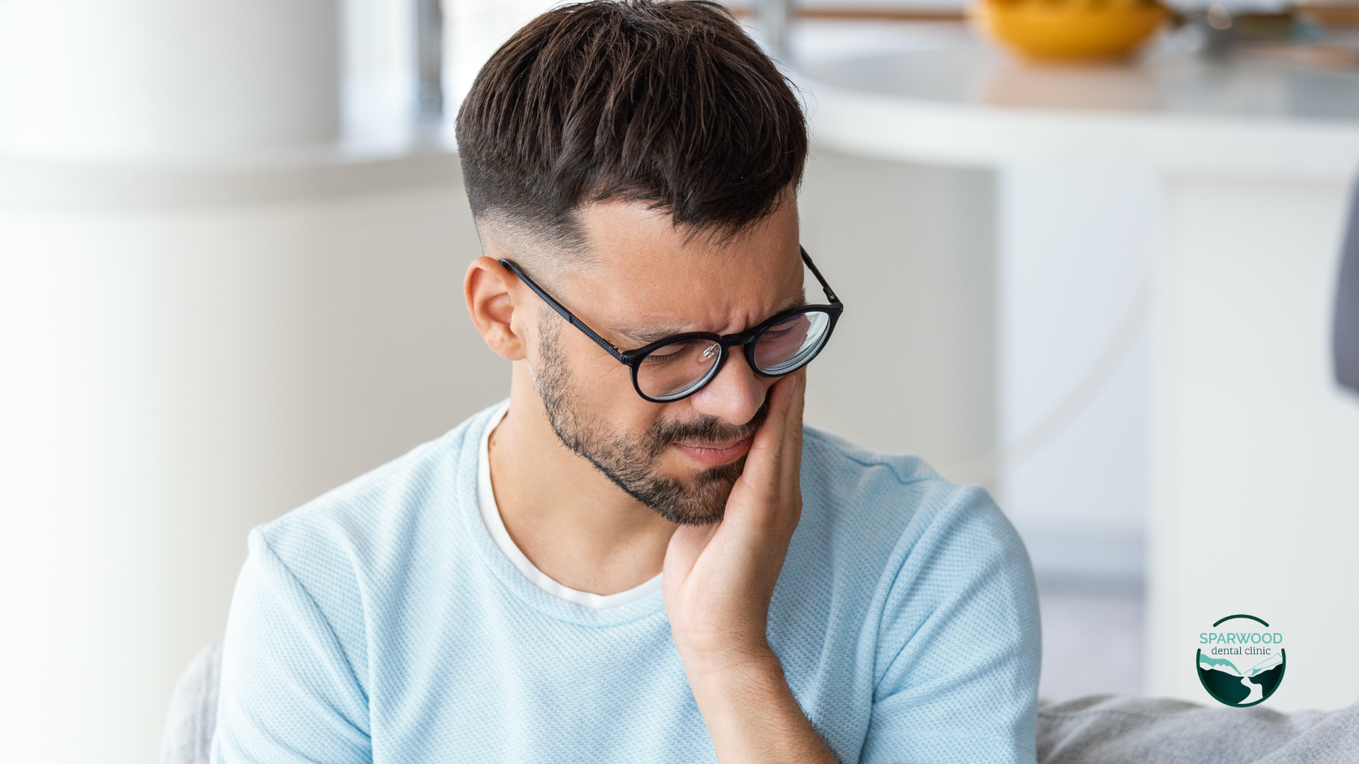 A man wearing glasses is sitting on a couch with a toothache.