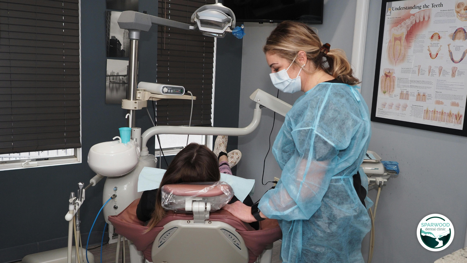 Dentist in blue gown and mask examining a patient in a dental chair. Clinic setting.