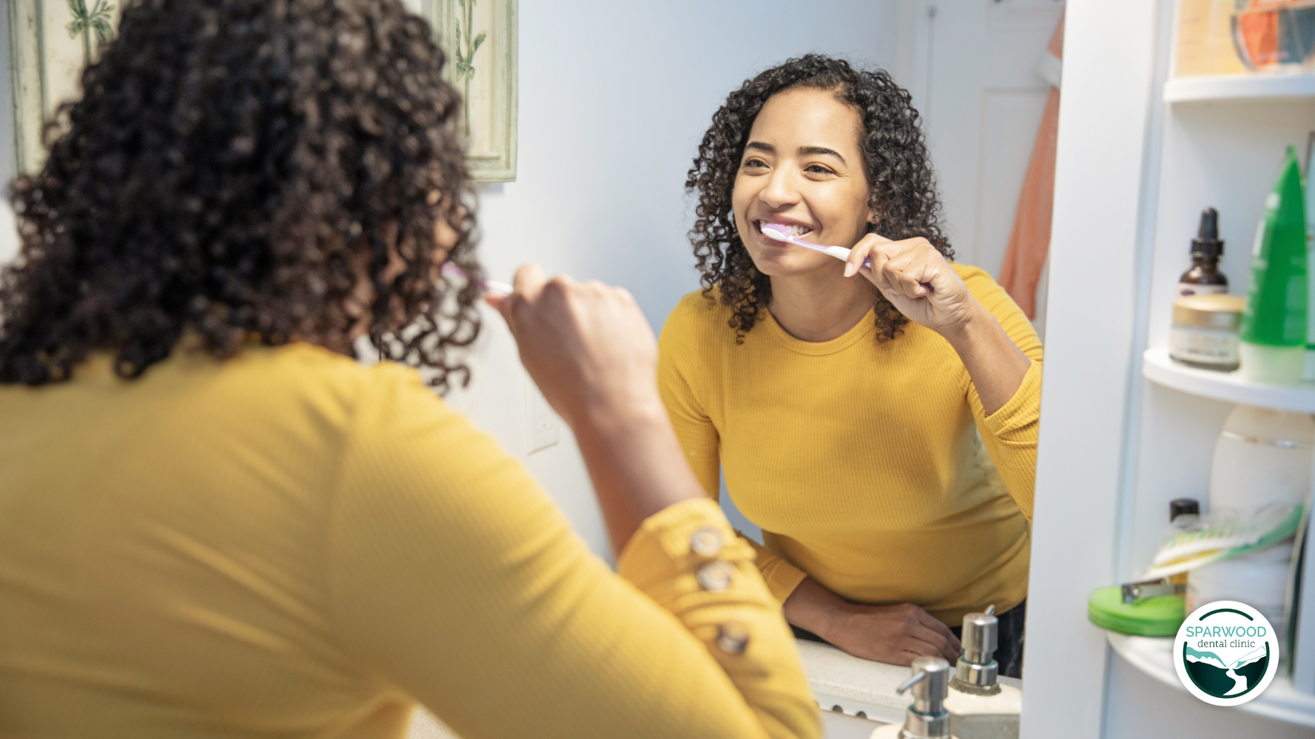 Woman brushing teeth in bathroom, yellow shirt, smiling.