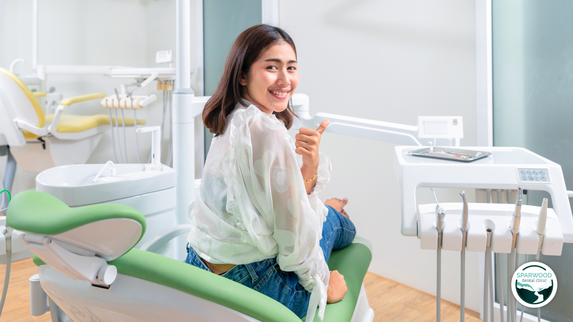 Woman smiling in a dental clinic, seated beside a green exam chair and dental equipment.