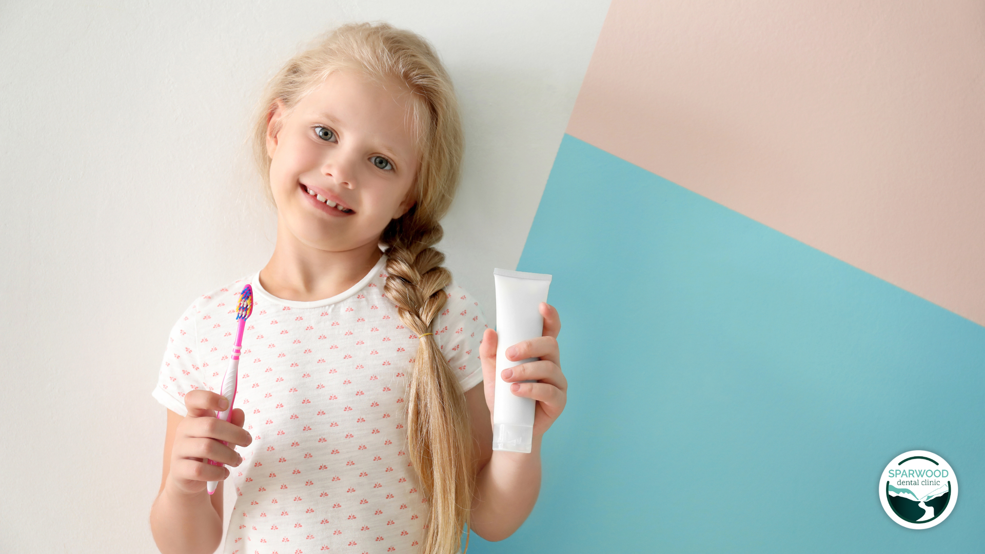 Girl holding toothbrush and toothpaste, smiling at the camera, leaning against pink and blue wall.