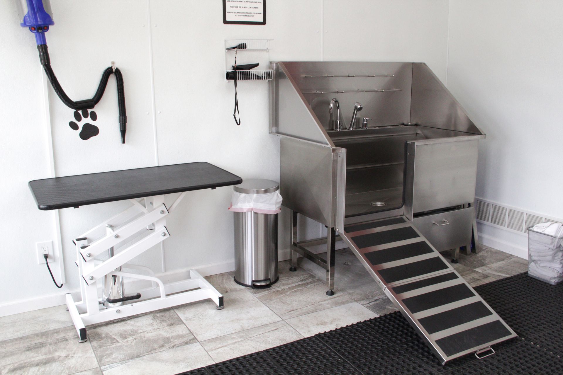 Grooming station: stainless steel dog wash with ramp, grooming table, and dryer in a bright room.