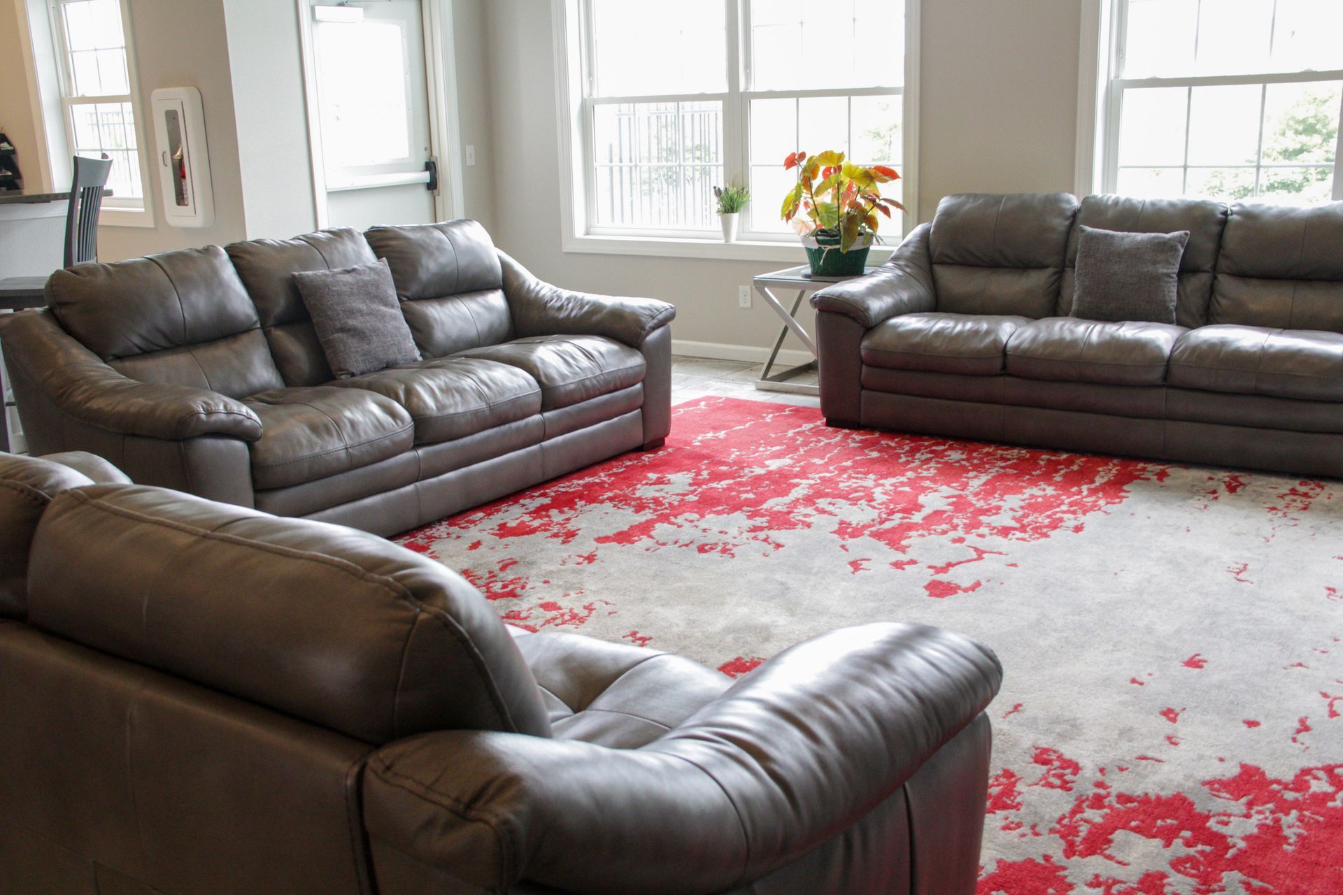 Living room with three brown leather sofas on a red and white rug, windows in the background.