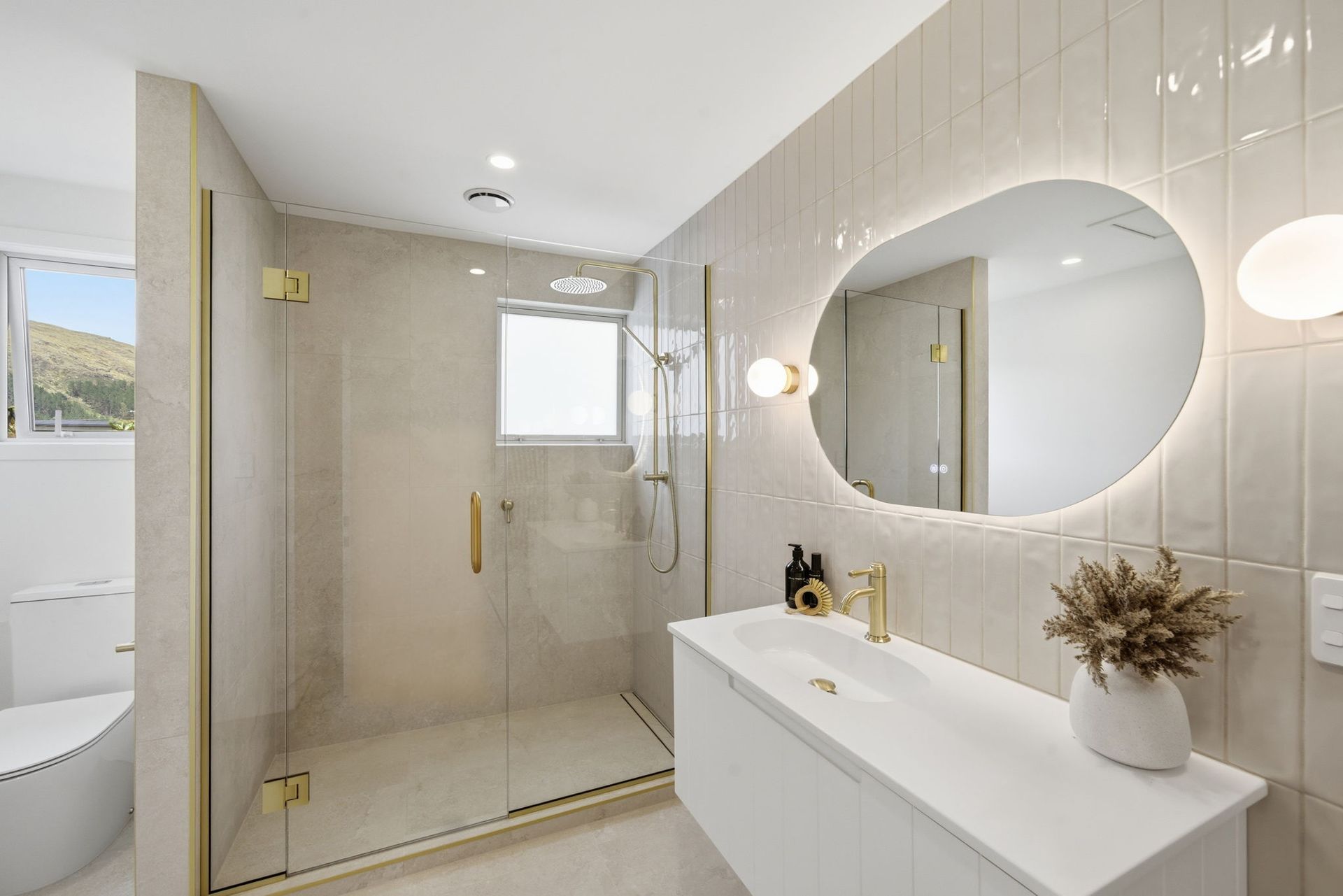 Modern beige bathroom with glass shower, round mirror, vanity, and large window
