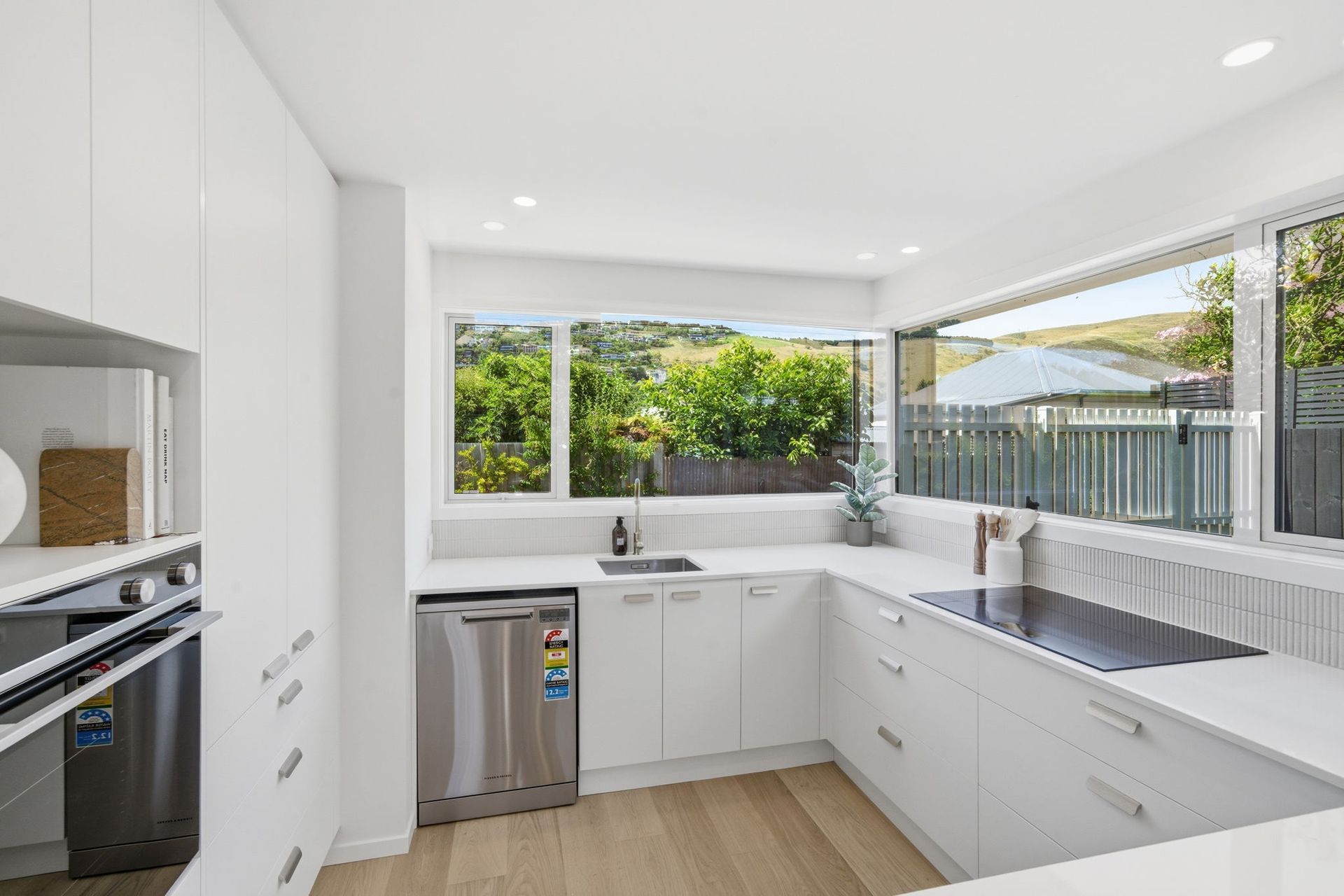 Modern white kitchen with large windows, stainless appliances, and light wood flooring