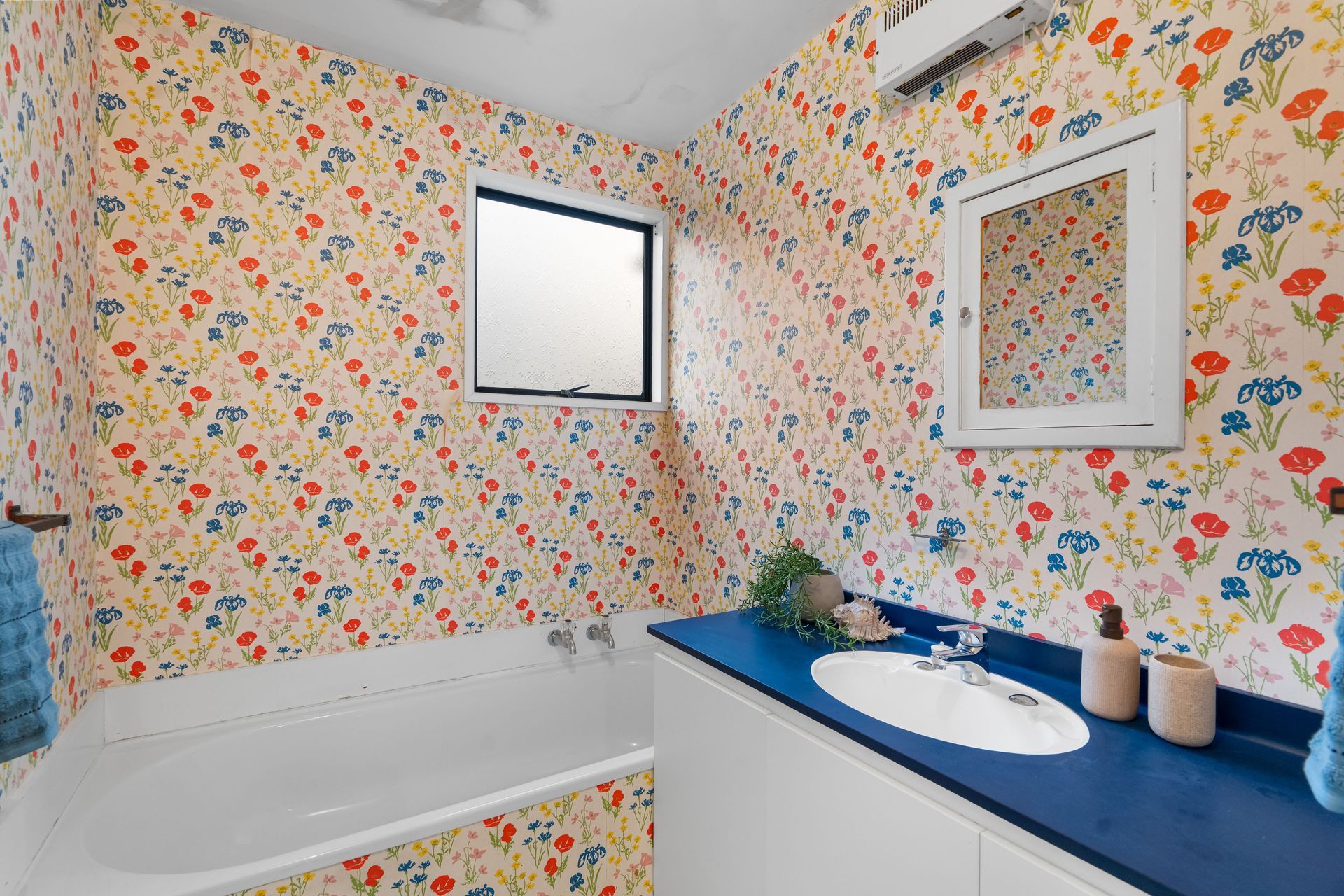 Colorful floral bathroom with a white tub, blue countertop, sink, mirror, and small window