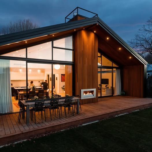 Modern wooden cabin with large windows, lit patio, and dining table at dusk