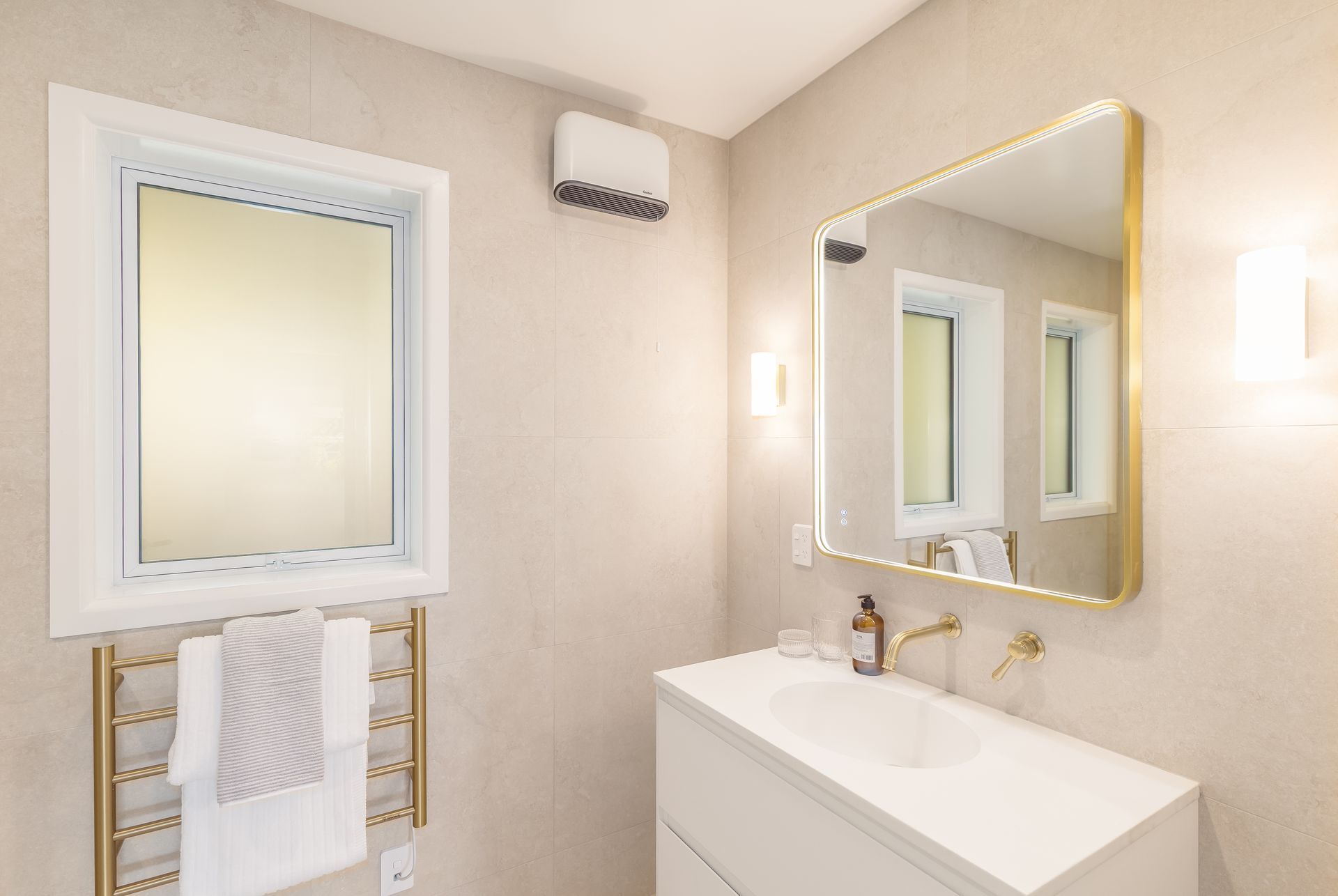 Bright bathroom with gold-trimmed mirror, white sink, wall lights, and towel rack near a frosted window