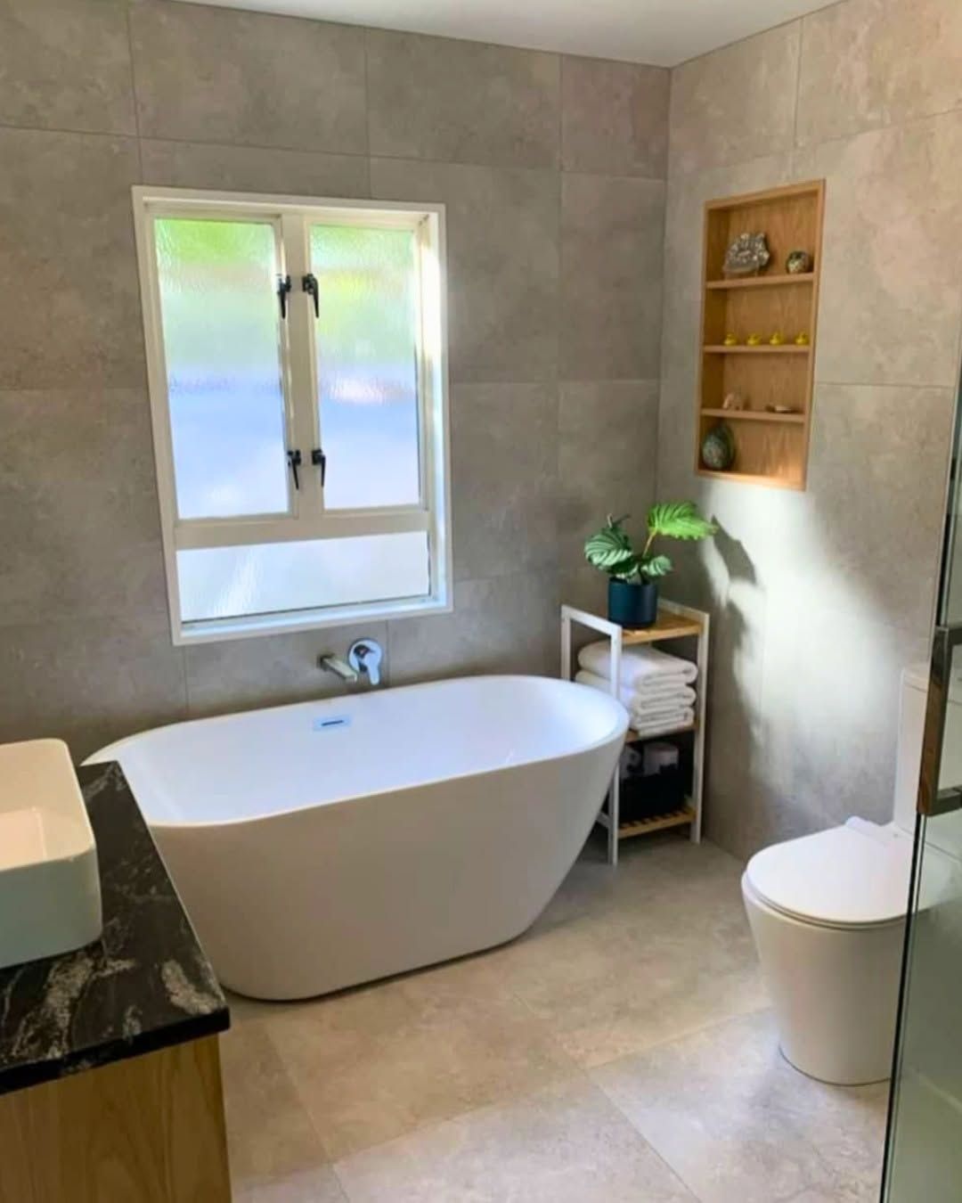 Modern bathroom with freestanding tub, window, toilet, and small shelf with plant