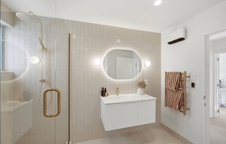 Modern bathroom with beige tile, floating vanity, oval mirror, glass shower, and wood towel ladder