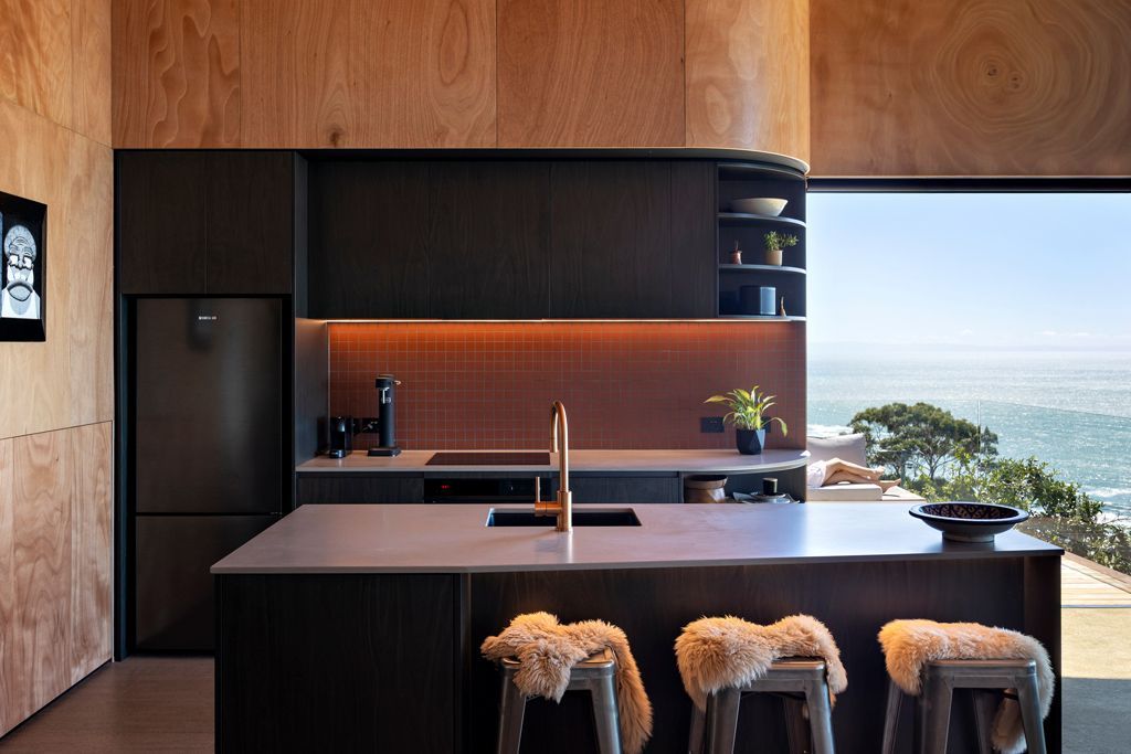 Modern kitchen with dark cabinetry, wood accents, island stools, and a large window overlooking the water