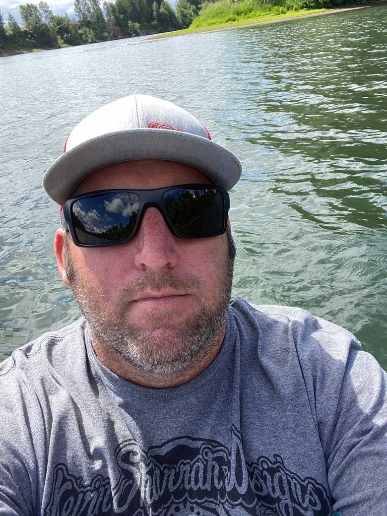 A man with a short beard wearing a grey cap and sunglasses takes a selfie while outdoors on a river.