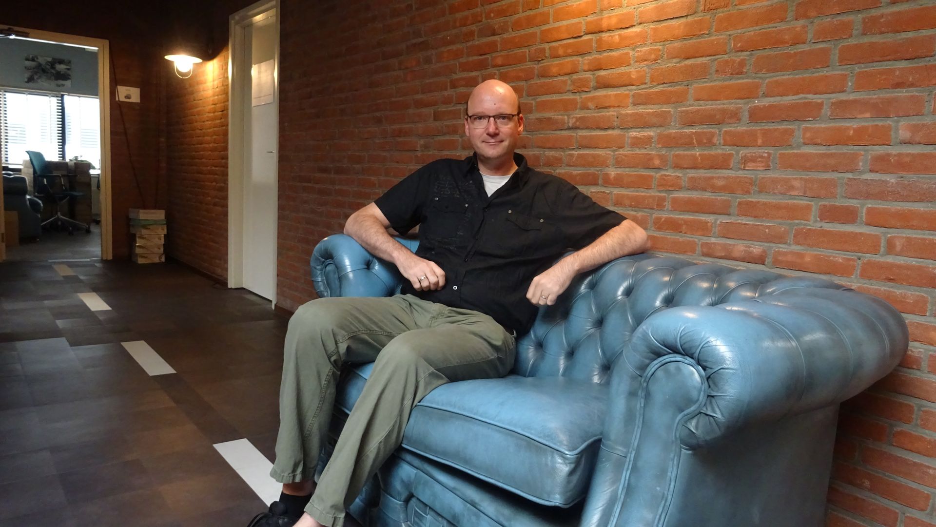 Een persoon zit op een blauwe, gecapitonneerde leren bank tegen een bakstenen muur in een kantoorgang.