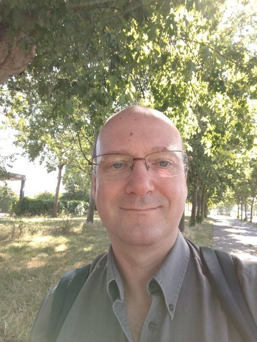 Een persoon met een bril glimlacht terwijl hij buiten staat op een pad dat omzoomd is met bomen.