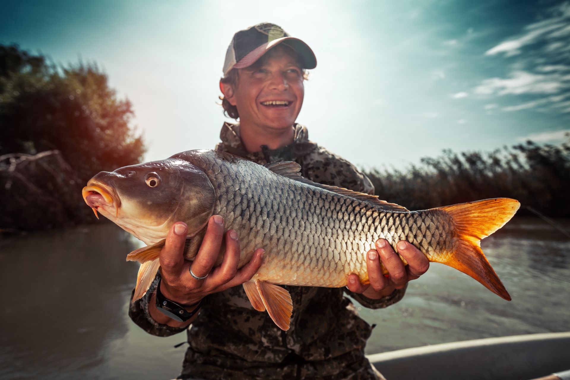 Man holding a large carp fish, smiling, near a river.